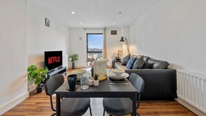 Moderni olohuone, jossa on ruokailutila, musta sohva, televisio, jossa näkyy Netflix, ja näkymä ulos.