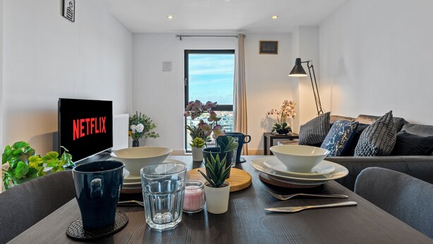 Moderni olohuone, jossa on Netflix-logo, ruokapöytä kulhoineen ja laseineen, sohva ja näkymä ovesta.