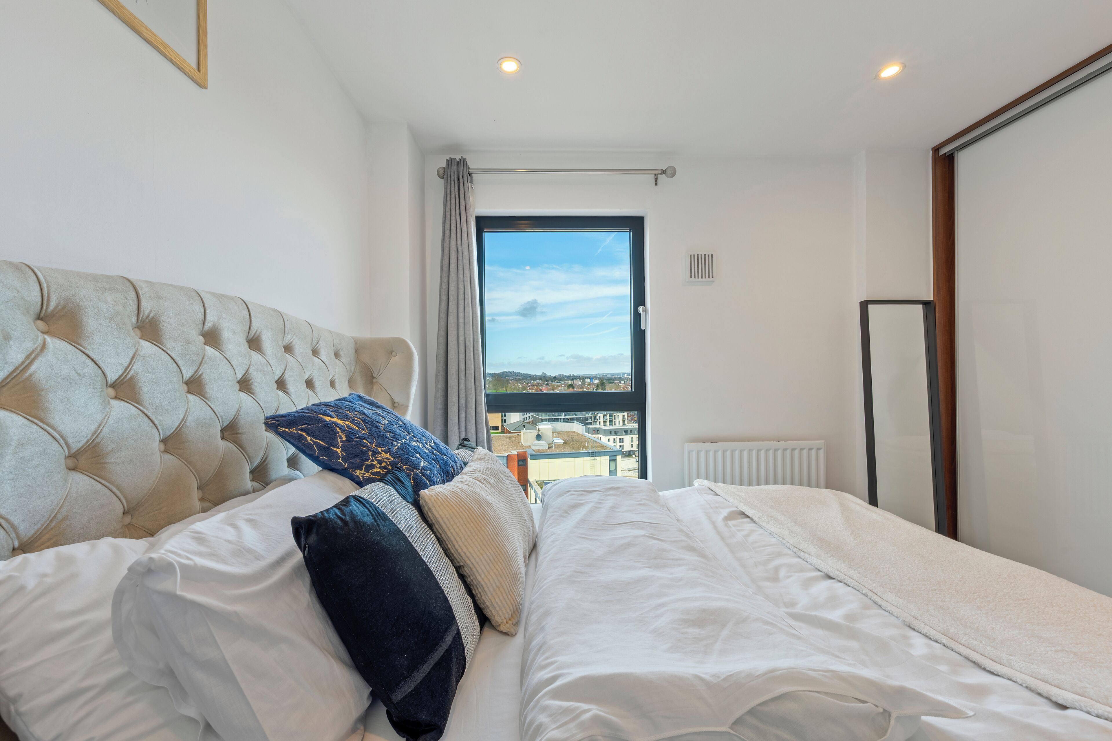 A bedroom with a tufted headboard, a large bed with white bedding, a view through a door to the outside, and a radiator on the wall.