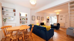 Una sala de estar con un sofá azul, una mesa de comedor de madera, sillas, una estantería y un televisor.