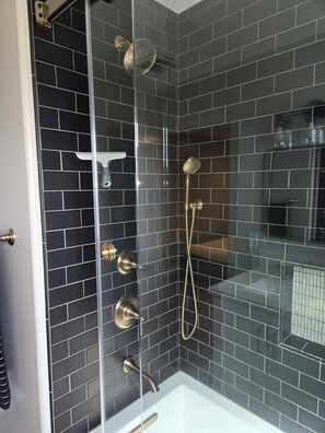 Una ducha moderna con cerramiento de vidrio, paredes de azulejos negros y una bañera blanca.