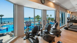 Une salle de sport équipée de machines de cardio, avec une horloge et une vue sur une piscine et une plage.