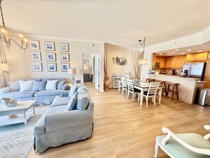 A spacious living room with a large sofa, a dining area with a kitchen in the background, and a hallway leading to another room.
