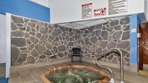 A spa area with a stone wall, a hot tub, a chair, and a metal safety rail. - Above the Clouds Views, Fireplace, Near Ski Resort (Beech Mountain)