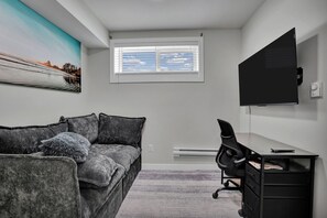 A room with a gray couch, a black office chair, a desk, a flat-screen TV, and a window with blinds.