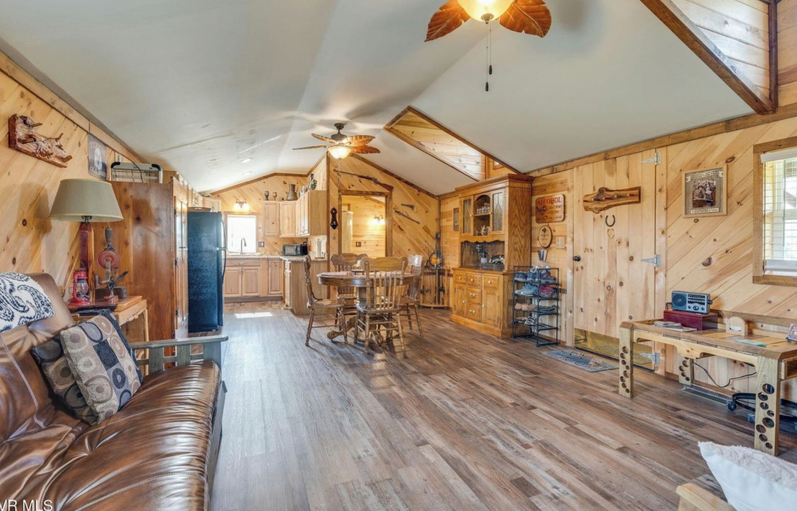 A cozy cabin interior with a wooden floor, a dining area, a kitchen, and a living room with a leather sofa.