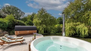 Un jacuzzi rodeado de sillas de descanso, una terraza de madera y un río con sauces.