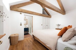Une chambre avec un plafond en pente, un lit avec des oreillers, une table de chevet, un miroir, une porte donnant sur une salle de bain et un panier en osier.