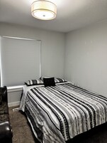 Un dormitorio con cama, mesita de noche, lámpara, ventana con persianas y una silla.