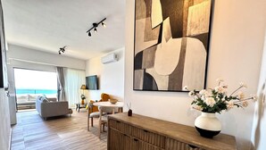 A modern hotel room with a wooden console table, a large abstract painting, a vase with flowers, and a view of the ocean through the window. - LuxuryOceanView Apartment | 16th Floor | 180°Views (Santo Domingo Este)