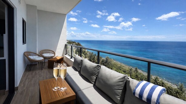 A balcony with a view of the ocean, a sofa, two wicker chairs, and a table with glasses. - LuxuryOceanView Apartment | 16th Floor | 180°Views (Santo Domingo Este)