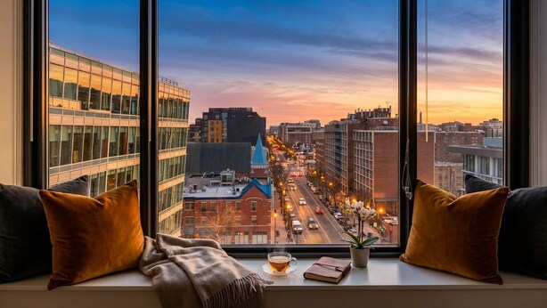 Une vue sur la ville, prise depuis une fenêtre, avec un canapé et des coussins, une tasse de thé et un livre posé sur le rebord de la fenêtre.