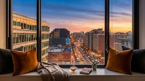 Vista panorámica de la ciudad desde una ventana con un sofá, cojines, una taza de té y un libro en el alféizar.