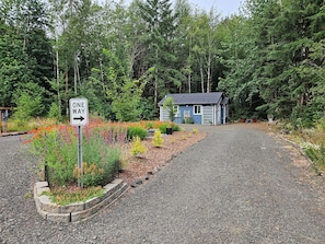 A gravel driveway leading to a blue building with a number 107 sign, surrounded by a garden with colorful flowers and dense forest in the background.