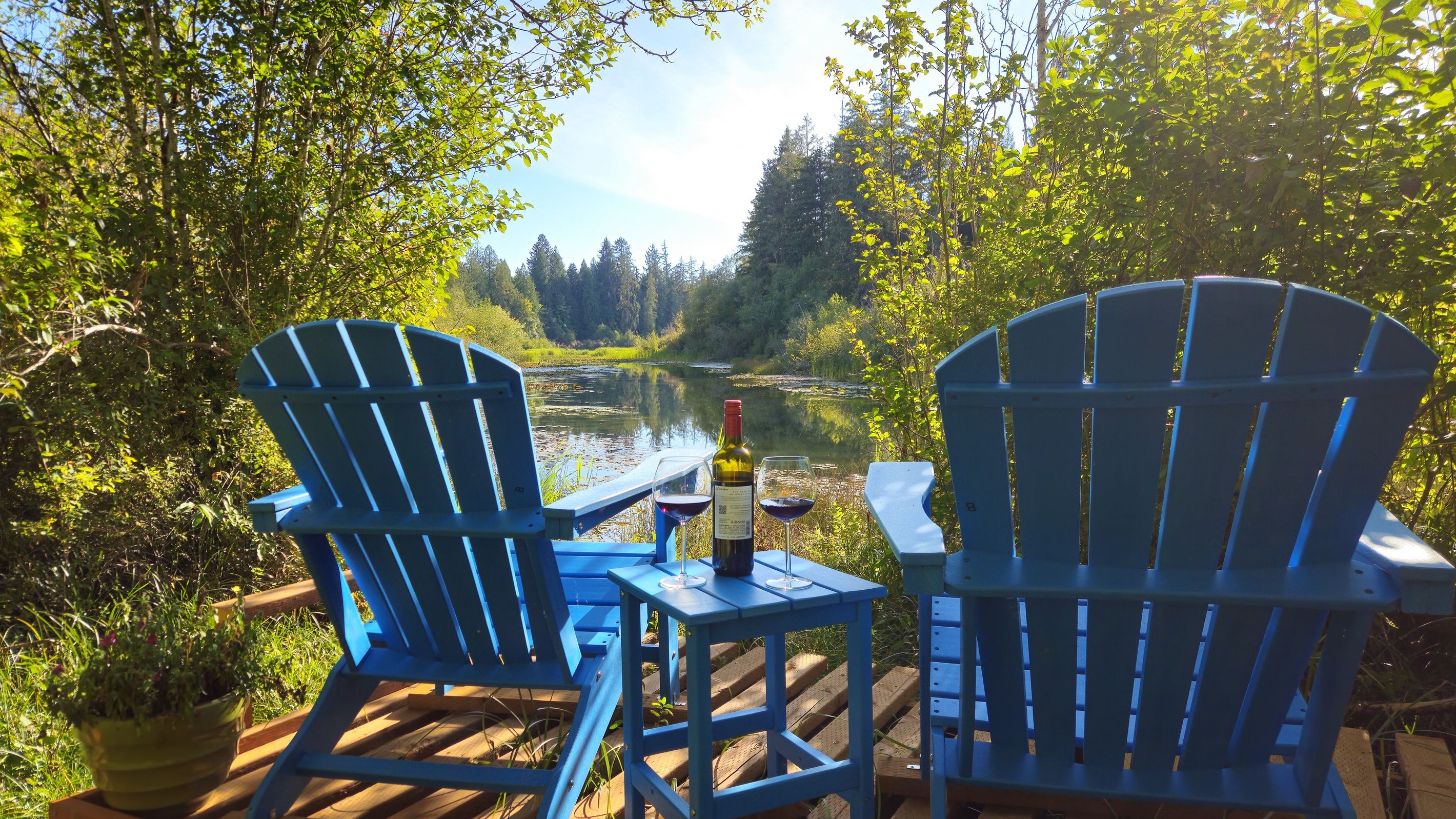 Two blue Adirondack chairs on a wooden deck overlooking a river, with a bottle of wine and glasses on the table.