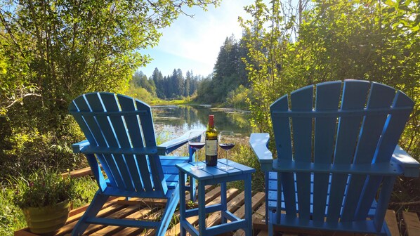 Two blue Adirondack chairs on a wooden deck overlooking a river, with a bottle of wine and glasses on the table.