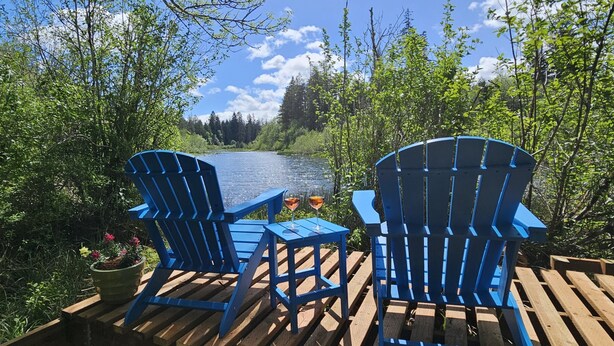 Two blue Adirondack chairs on a wooden deck overlooking a lake with trees and a clear sky.