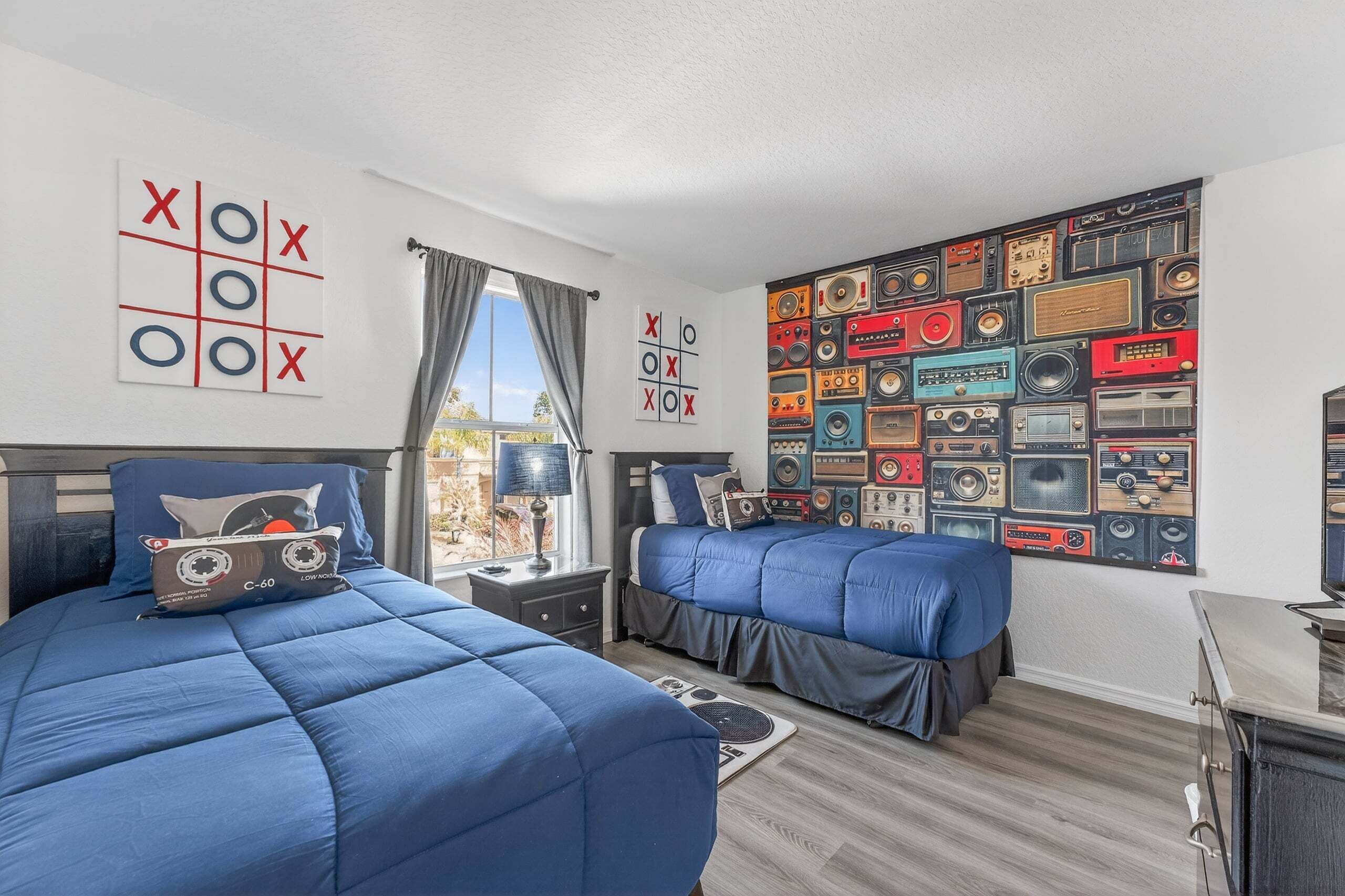 A room with two beds, a wall with vintage radio tapes, and a window with curtains.