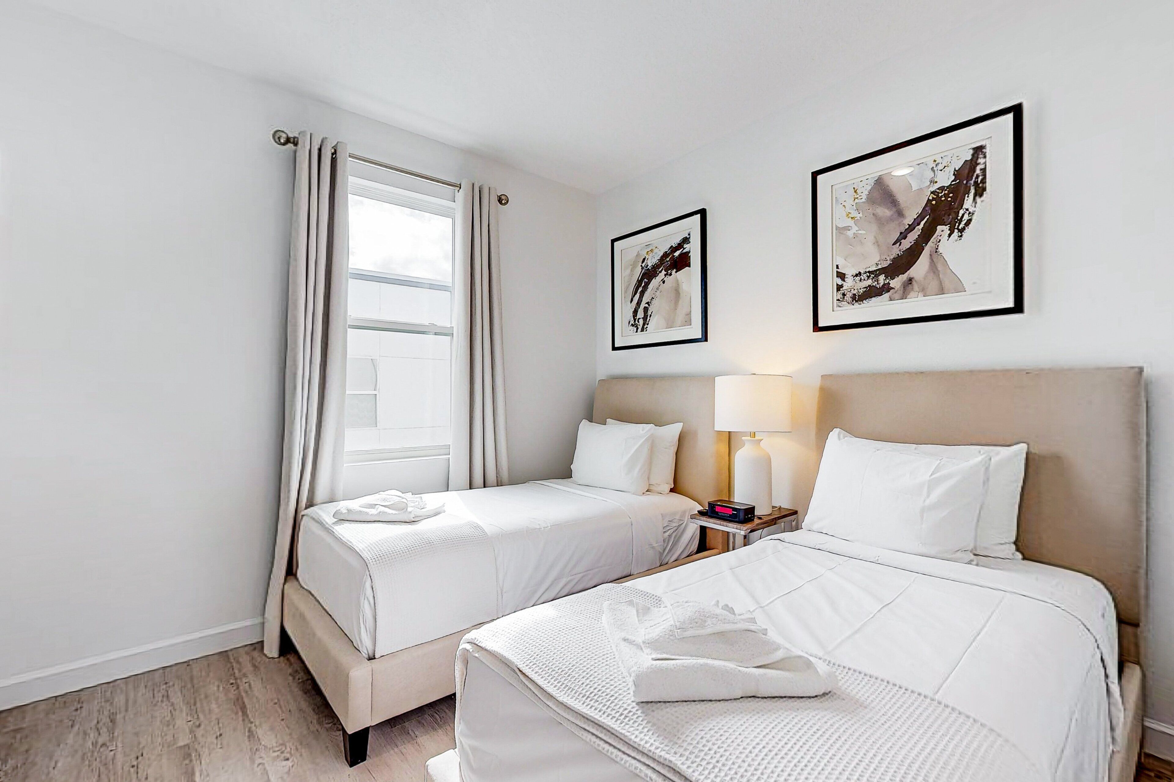 A hotel room with two beds, a window with curtains, a bedside lamp, and two framed artworks on the wall.