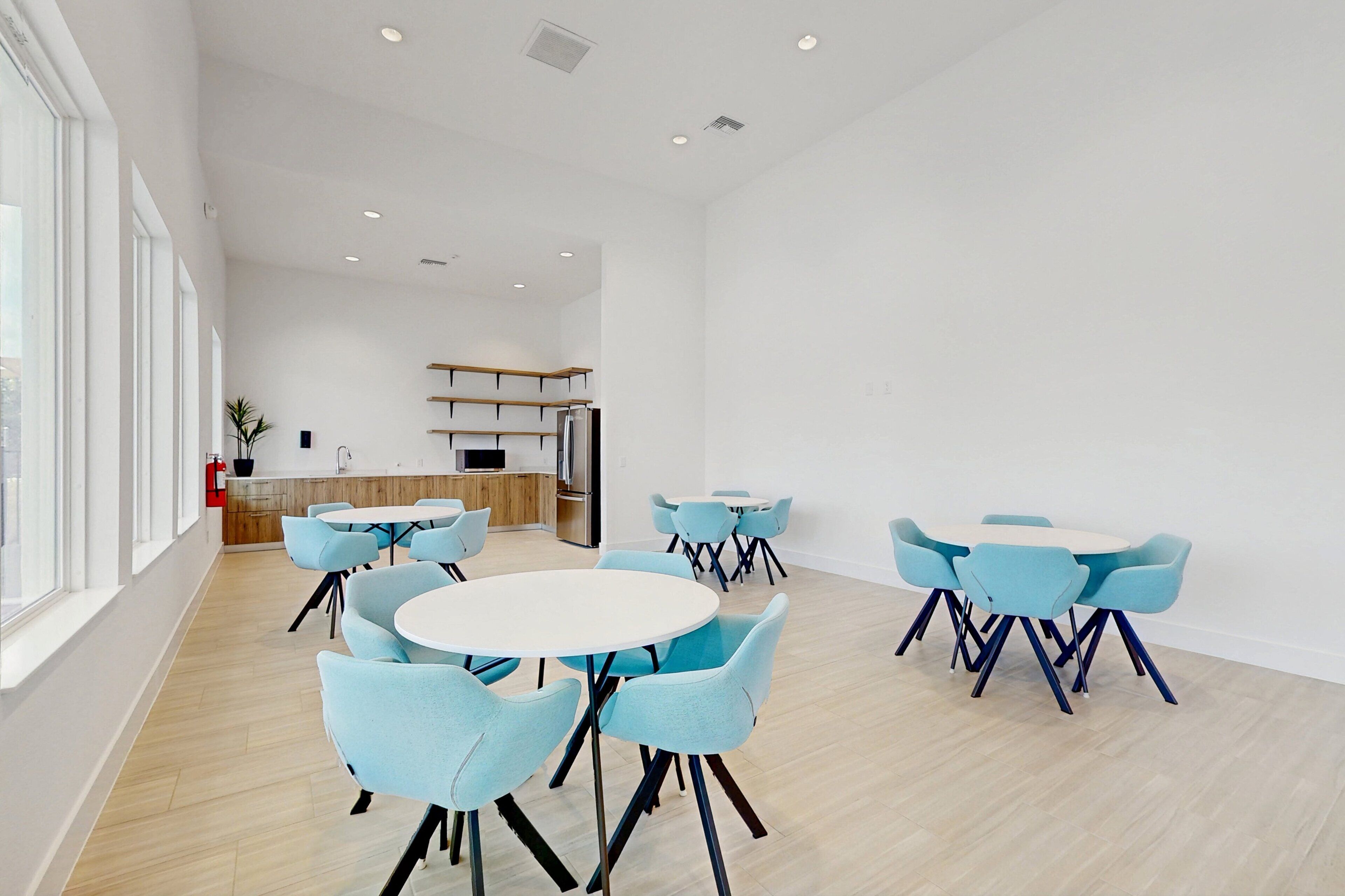 A modern dining area with round tables and blue chairs, a kitchen in the background, and a large window on the left.
