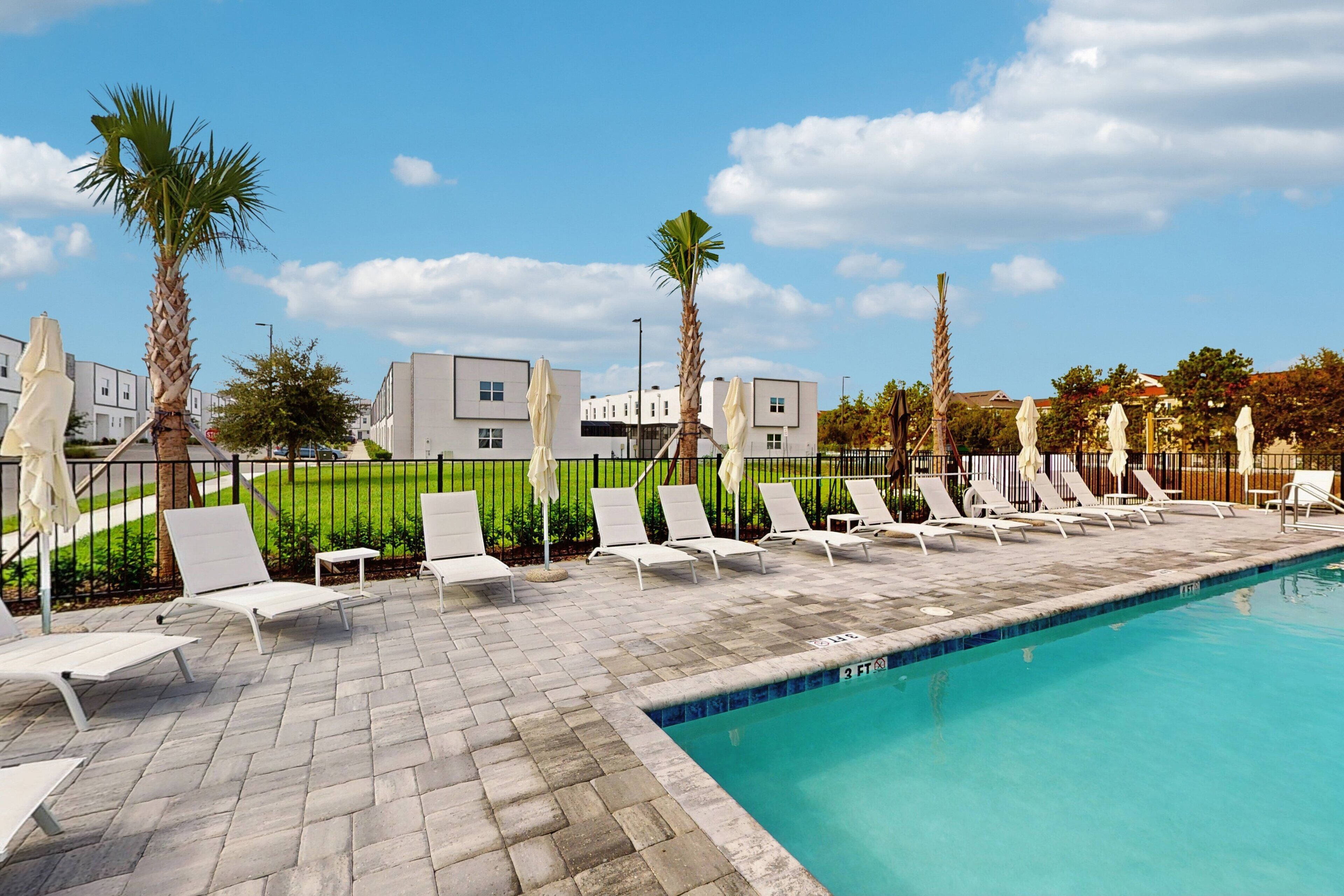 A pool area with lounge chairs and palm trees, surrounded by modern buildings.