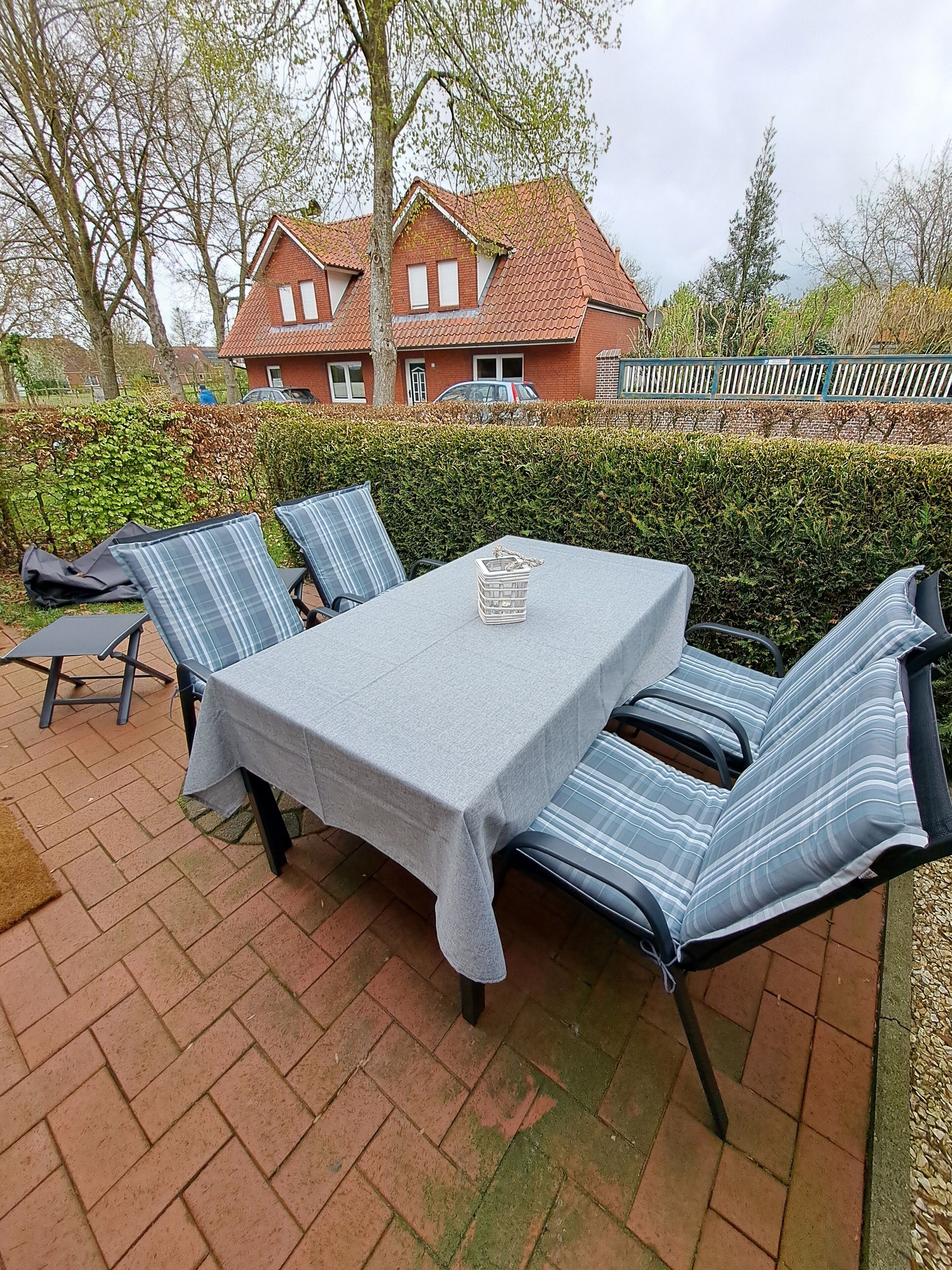 Een terras buiten met een tafel en stoelen, een haag en een huis op de achtergrond.