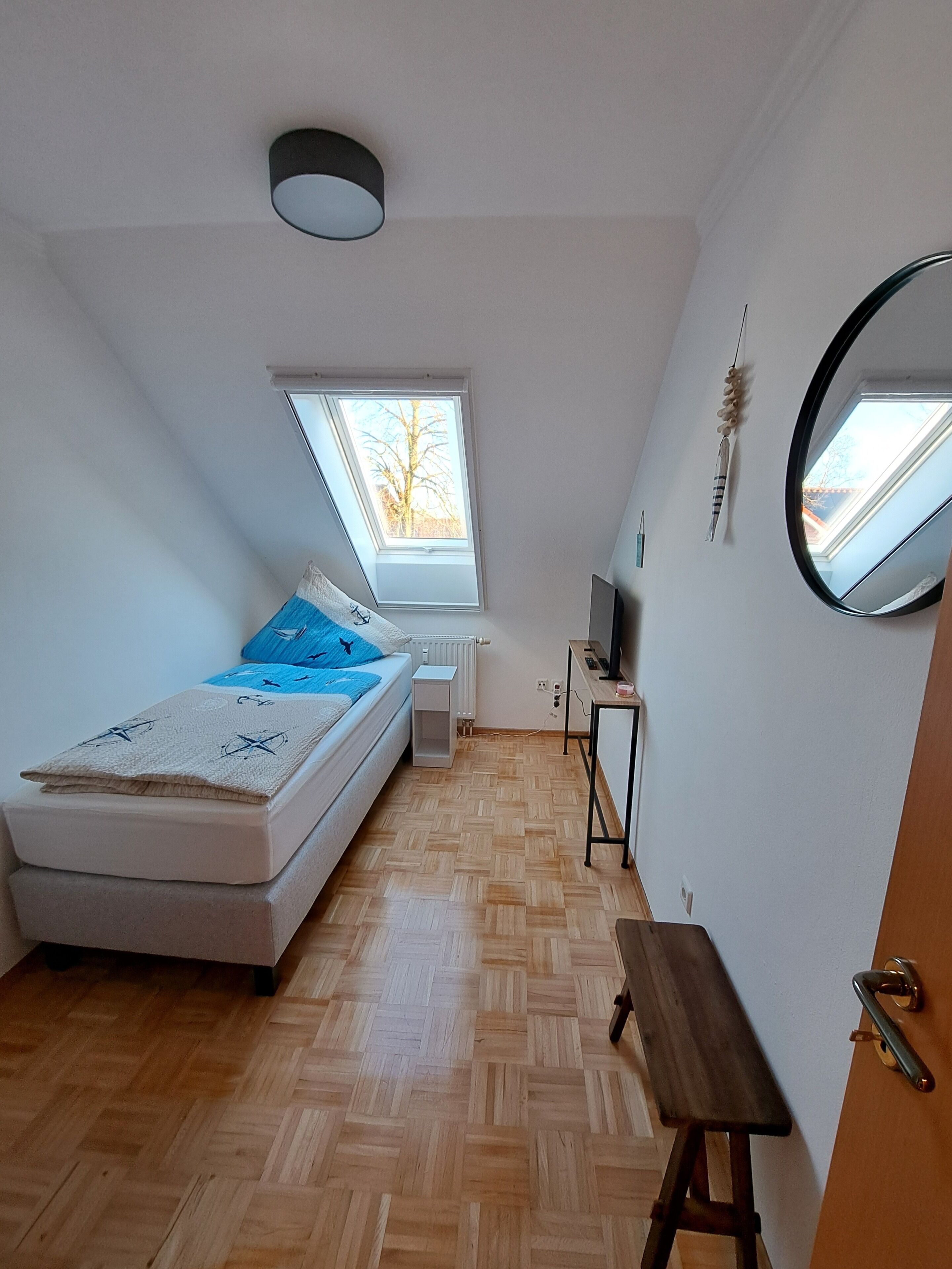 Un dormitorio con cama alta, un escritorio pequeño, un espejo y una ventana.