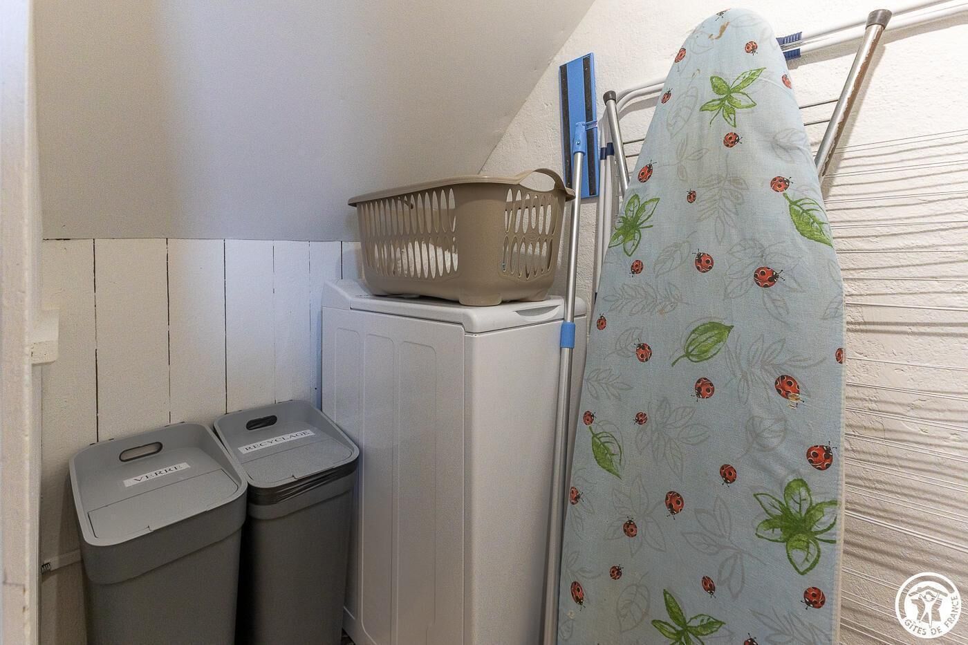 A laundry area with a washing machine, two trash bins, a drying rack, and a patterned shower curtain.