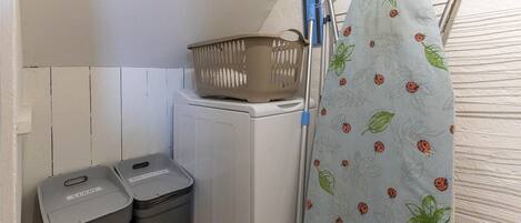 A laundry area with a washing machine, two trash bins, a drying rack, and a patterned shower curtain.