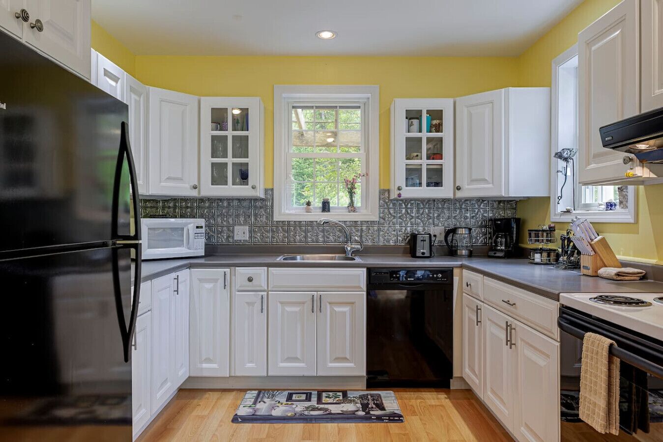 A kitchen with white cabinets, a stainless steel refrigerator, a microwave, a dishwasher, and a window with a view of greenery.