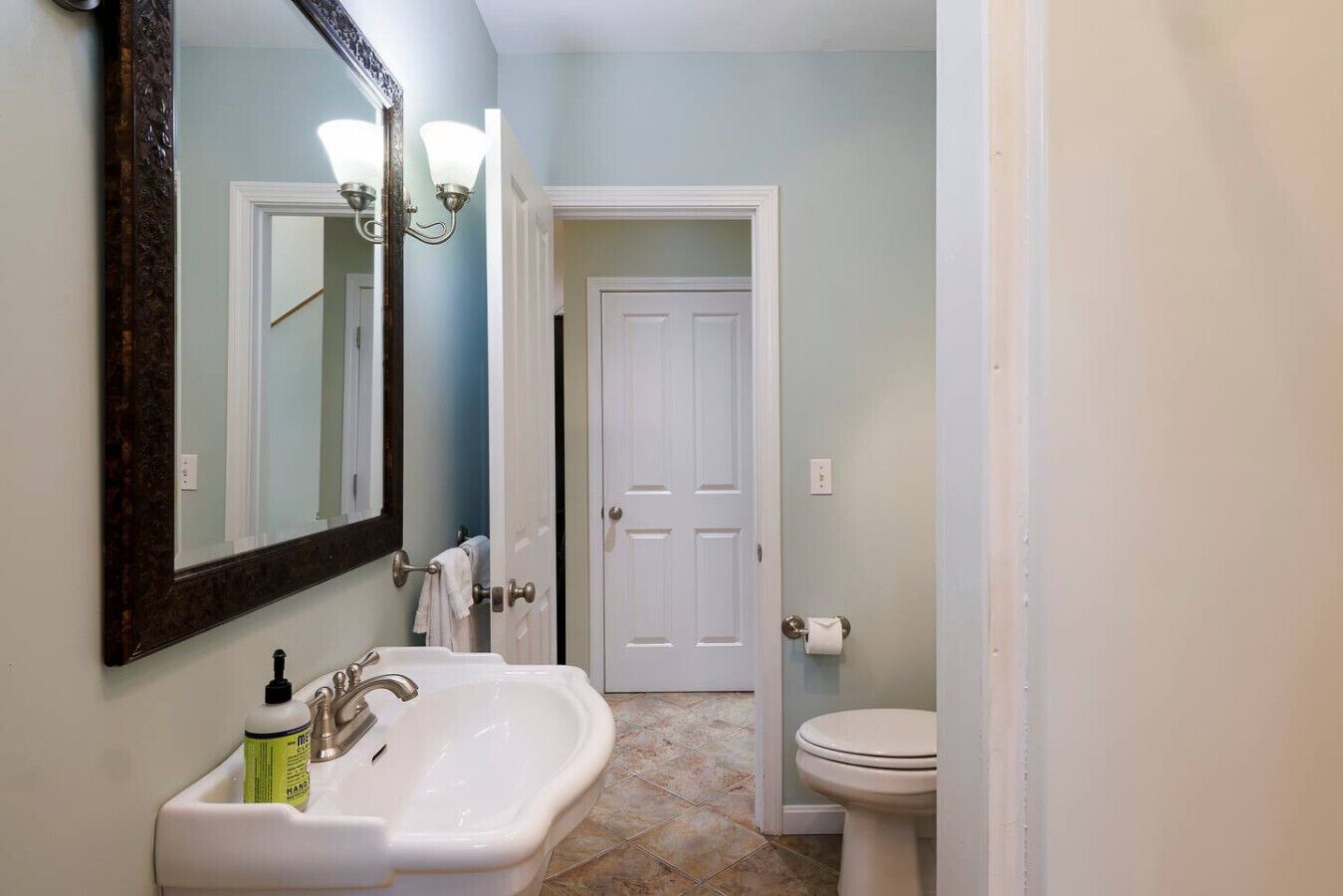 Une salle de bain avec un lavabo blanc, un miroir, des toilettes et une porte donnant sur une autre pièce.