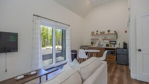 En moderne stue med sofa, fjernsyn, spisebord med stole, et køkkenområde med køleskab og en skydedør til terrassen.