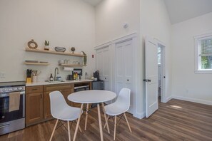 A modern kitchen with wooden cabinets, a built-in oven, a dining table with chairs, and a large window. - Quiet New Build Ellsworth Studio! [Solace Nook] (Ellsworth)