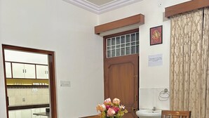 Uma sala de jantar com uma mesa de vidro, cadeiras de madeira, um vaso de flores, uma porta de madeira e uma cozinha visível através de uma porta aberta.