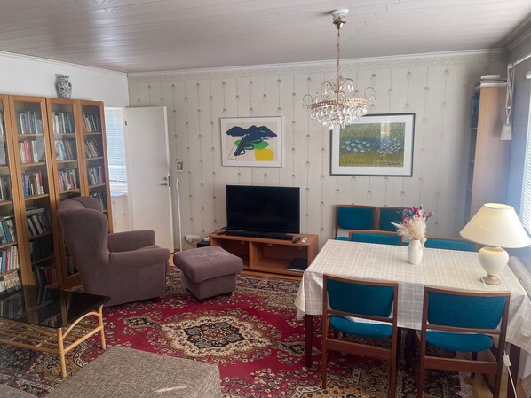Un salon comprenant un téléviseur, des étagères à livres, un lustre, une table à manger avec des chaises et un tapis à motifs.
