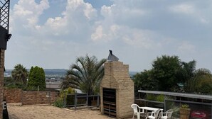 Um terraço no telhado com uma churrasqueira de pedra, cadeiras de plástico branco e vista para a paisagem urbana.