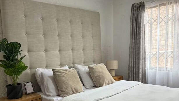 Quarto com cabeceira estofada, cama com roupa de cama branca, mesa de cabeceira de madeira com gavetas, planta em vaso e janela com cortinas.
