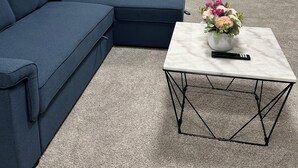 Un salón con un sofá seccional azul, una mesa de centro de mármol con un jarrón de flores y un control remoto.