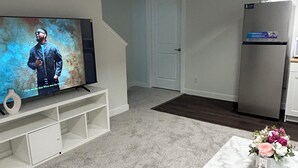 Una sala de estar con una televisión de pantalla plana, un mueble blanco para equipos electrónicos, una mesa de centro de mármol con flores, un refrigerador y una puerta que da a otra habitación.