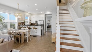 A modern kitchen with white cabinets, a dining area with a wooden table and chairs, and a staircase with wooden steps. - 3BR, 3BA, Villa steps from sand with private beach access, beach gear & pool! (Miramar Beach)