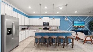 A modern kitchen with white cabinets, a stainless steel refrigerator, a central island with stools, pendant lighting, and a blue accent wall. - Upstay Disney Family Adventure with Pool & Hot Tub (Davenport)