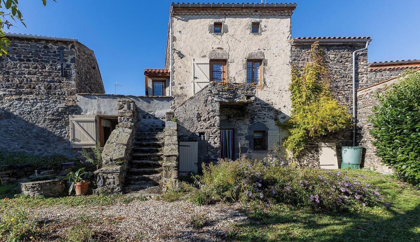 Un bâtiment en pierre avec un escalier menant à une porte, entouré de verdure et sous un ciel dégagé.