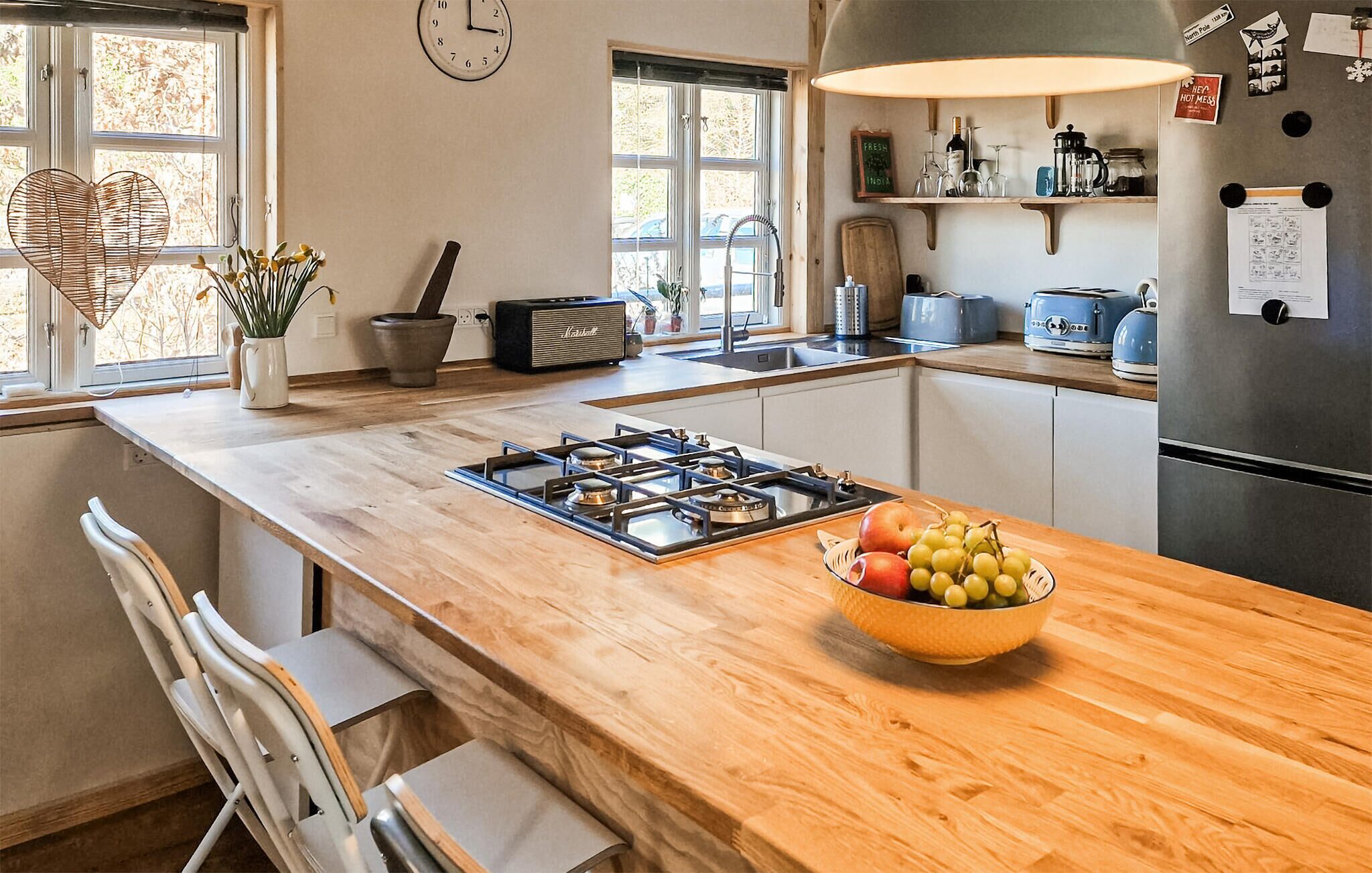 A kitchen with a gas stove, a bowl of fruit, a clock, and a window with a view of trees.