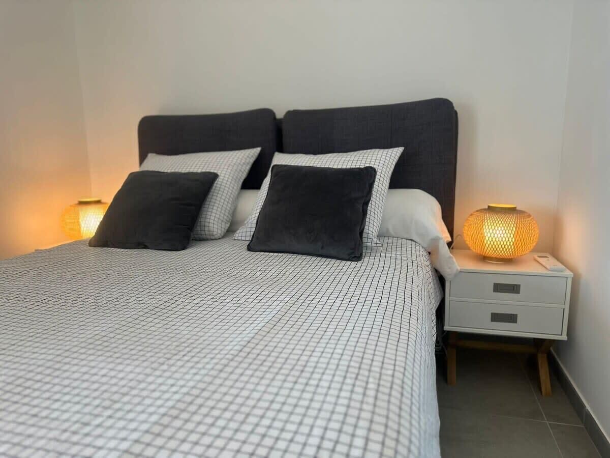 A bed with a checkered bedspread, two black pillows, a bedside lamp, and a nightstand with drawers.