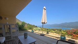 Eine Holzterrasse mit Gartenmöbeln, einem Tisch und einem Sonnenschirm mit Blick auf eine malerische Landschaft aus Meer und Bergen.