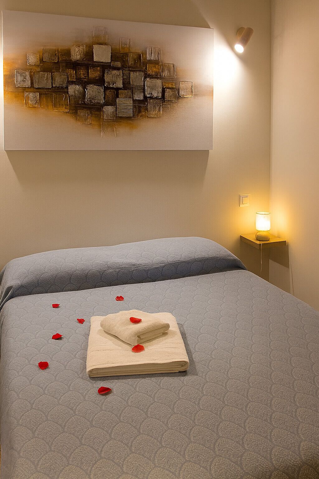 Une chambre d’hôtel avec un lit, une œuvre d’art accrochée au mur, une lampe de chevet et une serviette soigneusement pliée, ornée de pétales de rose.