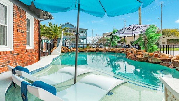 A swimming pool with lounge chairs and a slide, surrounded by a brick wall and palm trees. - Charmed Lagoon (Galveston)