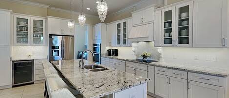 Una cocina moderna con armarios blancos, encimeras de granito, una isla central, electrodomésticos de acero inoxidable e iluminación colgante.