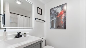 Een moderne badkamer met een wit wastafelmeubel, een spiegel, een toilet en een kunstwerk in een lijst aan de muur.