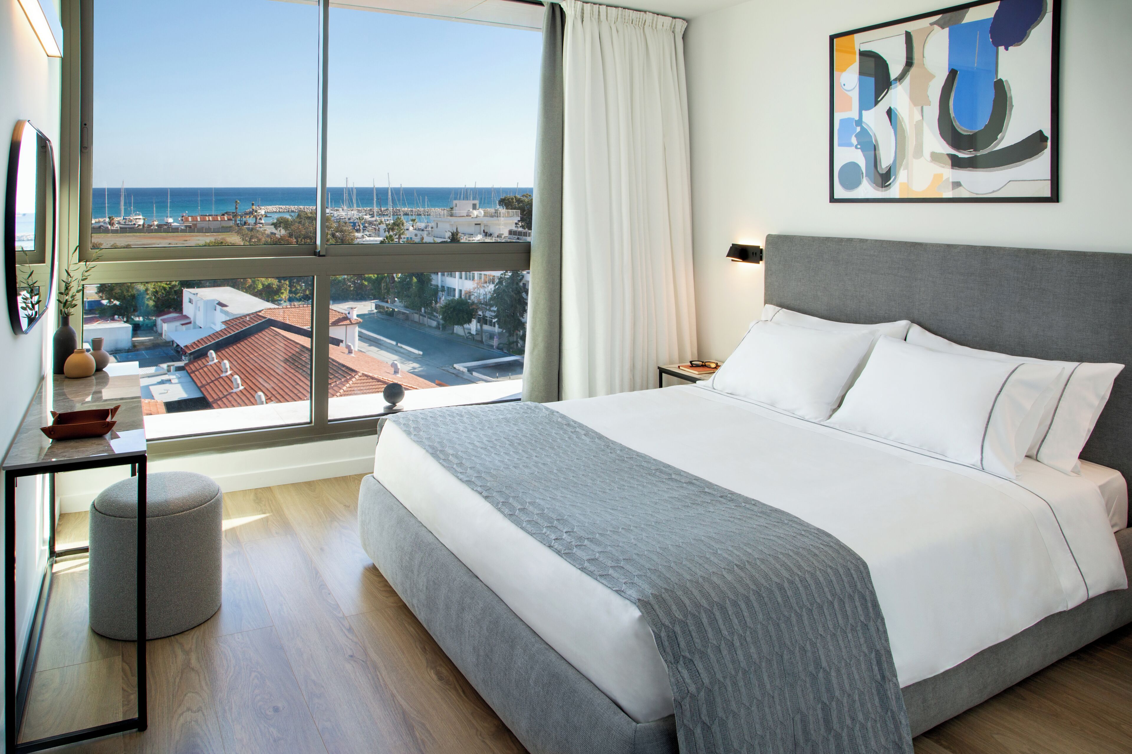 A bedroom with a large window, a bed, a bedside table, and a wall-mounted artwork.